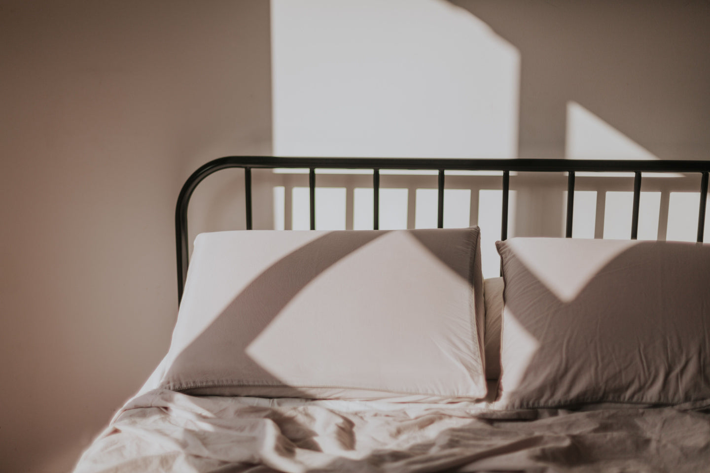 Opgemaakt bed met twee lichte kussens tegen een zwart spijlenhoofdbord in zonlicht