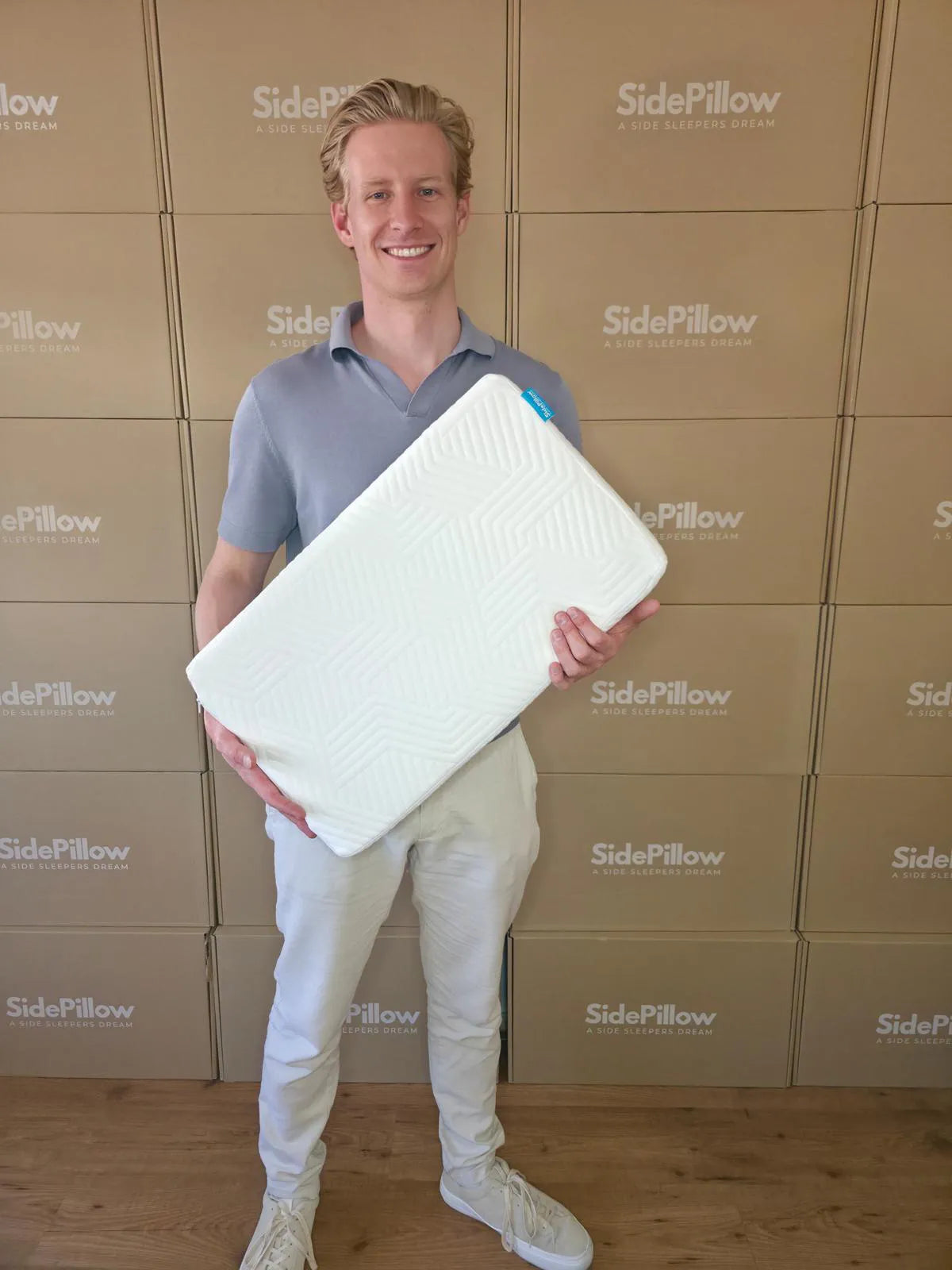 Oprichter Björn Braaksma houdt een rechthoekig SidePillow hoofdkussen vast voor een muur met SidePillow dozen.